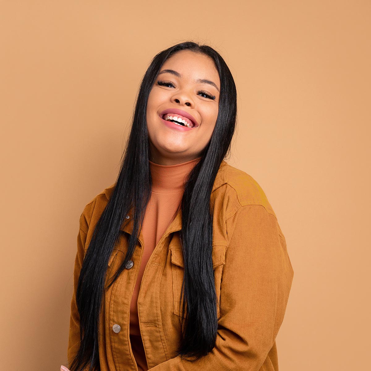 Smiling young woman with long black hair wearing a brown jacket, radiating confidence and happiness against a warm, neutral background.