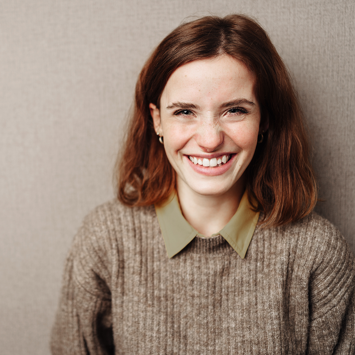 Smiling young woman with auburn hair wearing a cozy brown sweater and green collar, radiating warmth and happiness against a neutral background.