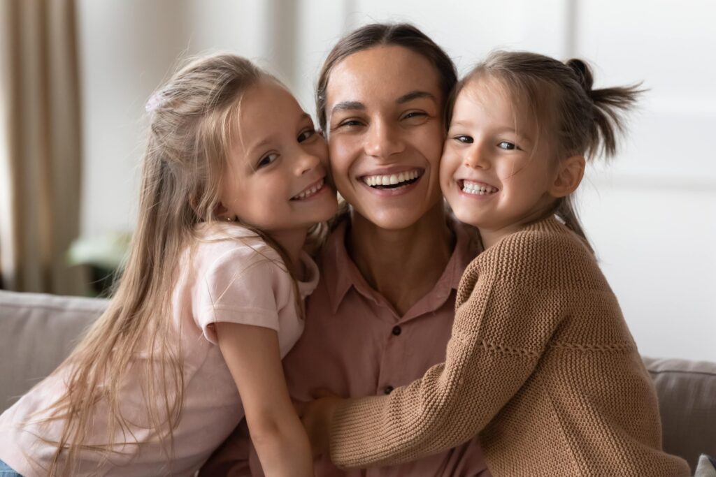 Smiling mother hugging her two young daughters, enjoying a close family moment in a cozy indoor setting.