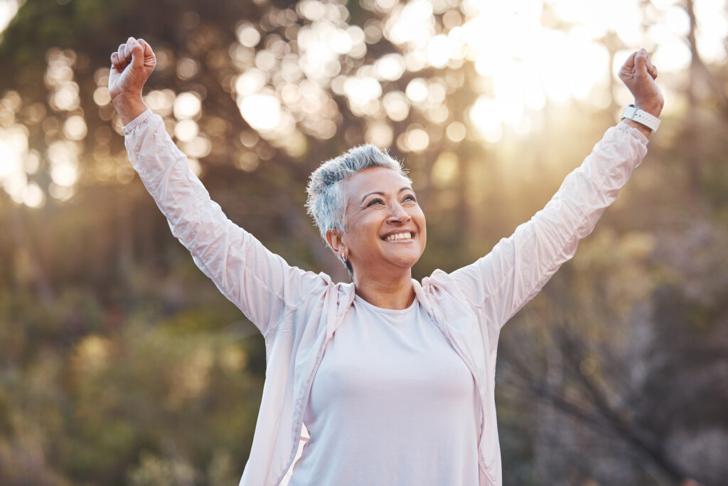 Happy senior woman outdoors with arms raised in celebration, enjoying a healthy, active lifestyle in a sunlit natural setting.