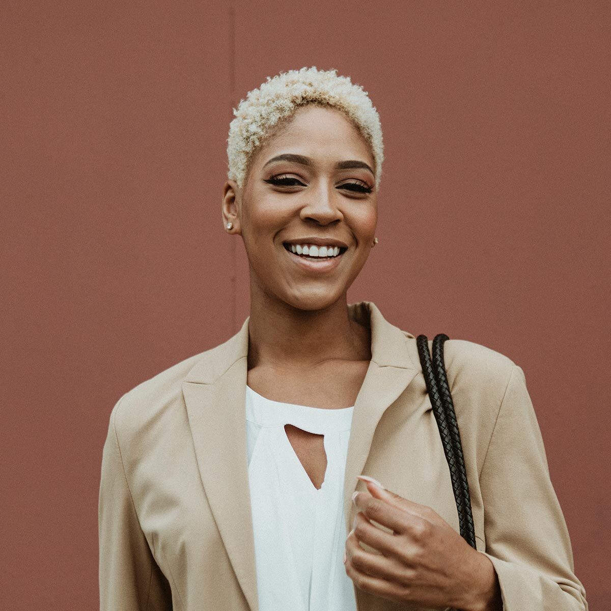 Confident young woman with short blonde hair smiling in a beige blazer and white top, standing against a reddish background.